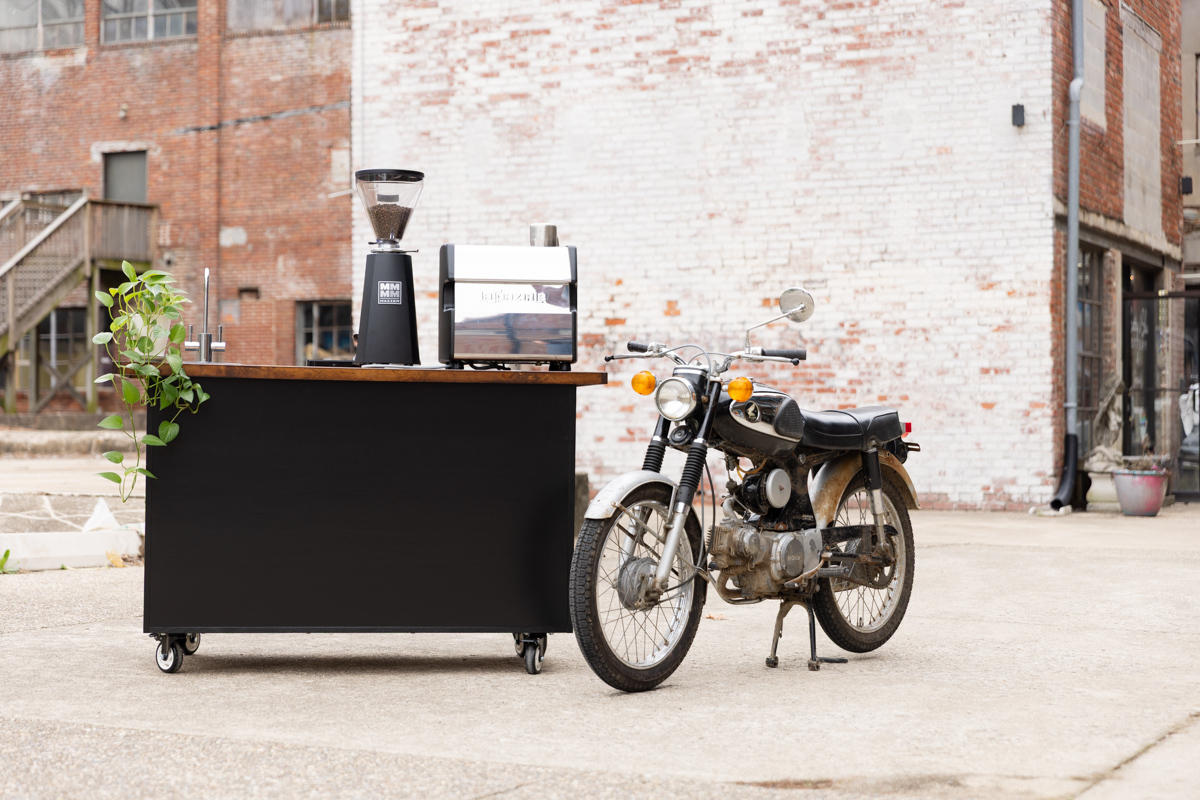 Black Kickstart Coffee Cart with a Nuova Simonelli LUCCA espresso machine and Mazzer grinder with a classic motorcycle sitting next to it.