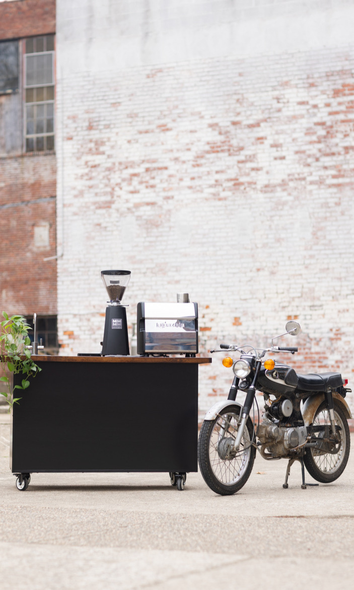 Black Kickstart Coffee Cart with a Nuova Simonelli LUCCA espresso machine and Mazzer grinder with a classic motorcycle sitting next to it.