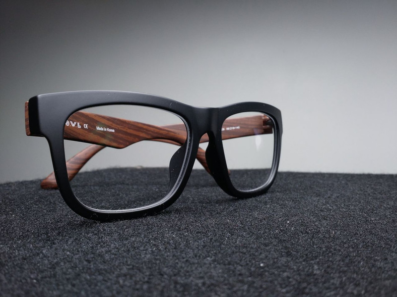Chunky matte-black square eyeglass frames with wood-finish temples on dark felt