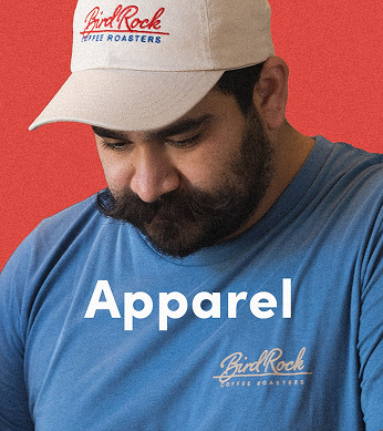 Male with mustache and beard wearing a tan hat and a blue shirt, both branded with Bird Rock Coffee Roasters: Apparel Shopping Category