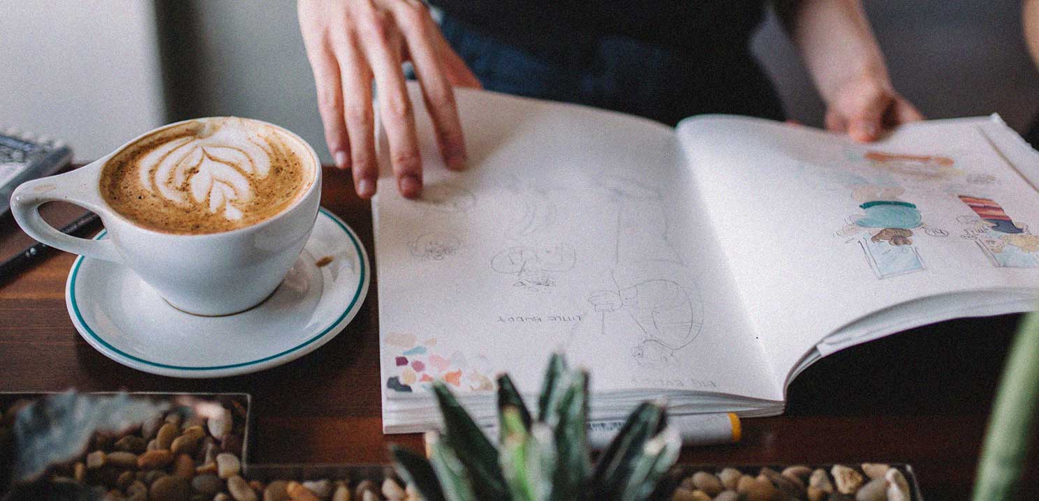 A latte with a design and a person looking at a book with various unknown hand drawn images.