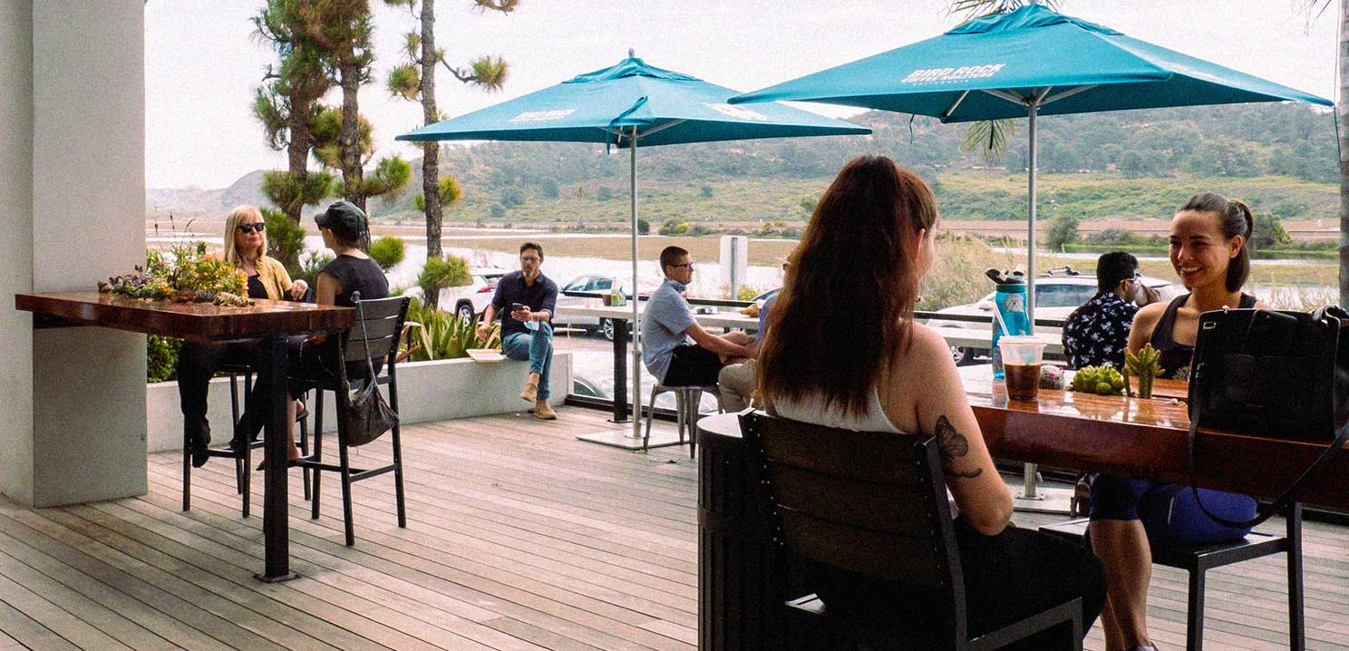 An outside patio at the Torrey Pines cafe showing multiple wood tables and blue umbrellas with people sitting in various locations.