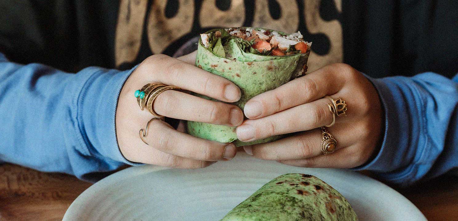 A person with blue sleeves holding a burrito or wrap with a green tortilla.