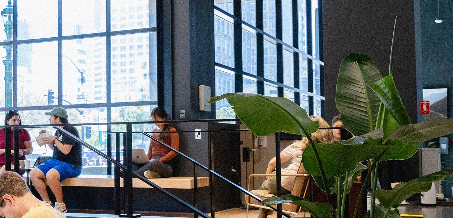 Interior of the Waterfront Cafe, showing people casually sitting in various areas in an industrial setting. There are with large windows and a plant as well.