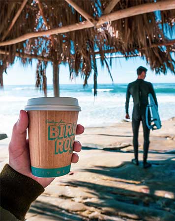 Hand holding a paper cup from Bird Rock Coffee with a surfer and the ocean in the background