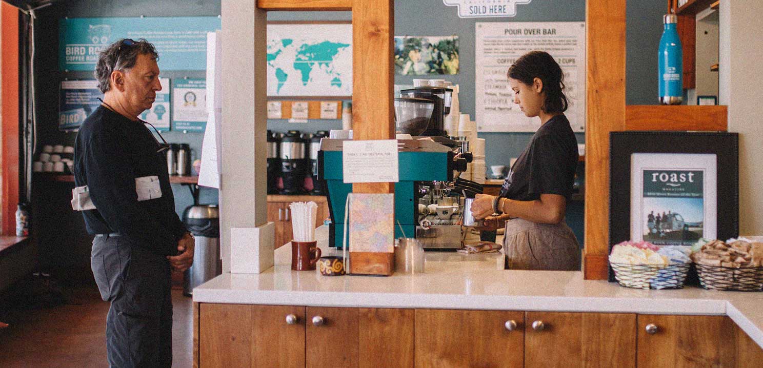 A man standing in front of a counter where a barista is preparing an espresso drink inside the La Jolla Cafe.