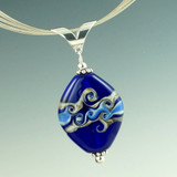 A beautiful base of deep cobalt blue with rich layers of ivory and blue glass with silver make this swirl pendant really pop. After shaping, the surface of one side is heated and then swirled with a thin rod of glass to create its own beautiful design, while the other side is left natural. Each each side offers its own distinct style.