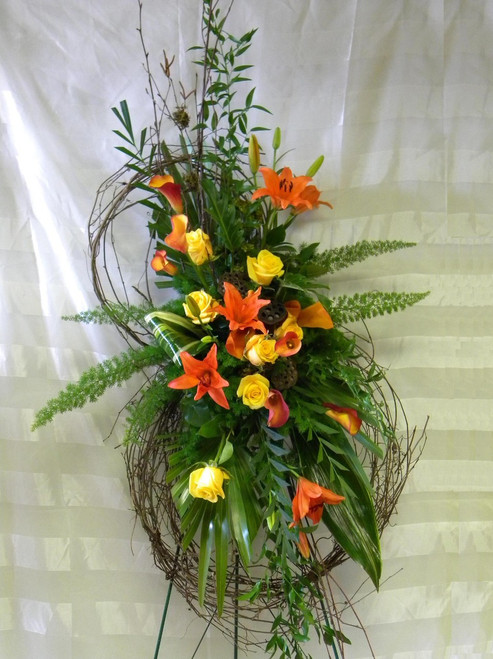 Autumn Harvest Grapevine Funeral Flower Wreath by Enchanted Florist TX. Send unique and creative funeral flowers with this grapevine flower wreath standing spray. A custom creation by our talented designers. Sympathy flowers include orange lilies, yellow roses, and orange mini calla lilies with tropical foliages arranged on this grapevine wreath. Approximately 70"H x 28"W. (size does not include stand) 
SKU RM519