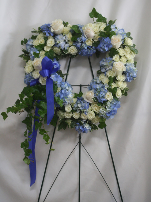 Blue Heart Funeral Tribute Flower Spray by Enchanted Florist Pasadena TX. Accented with trailing ivy, this sweet heart-shaped wreath of sky blue hydrangea, white big roses and pure white mini spray roses is a loving remembrance. SKU RM563