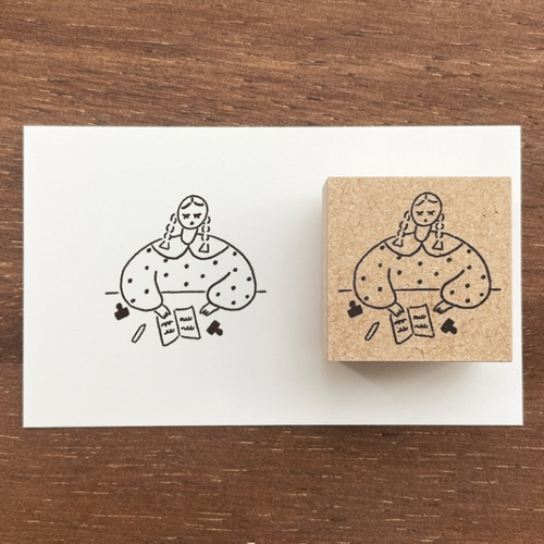 A square Japanese rubber stamp (a-089) featuring the character Marl-chan sitting and using a small rubber stamp on a piece of paper, celebrating the joy of stamping and crafting. Un tampon en caoutchouc japonais carré (a-089) illustrant le personnage Marl-chan assise en train d'utiliser un petit tampon sur une feuille de papier, célébrant la joie de l'estampage et de l'artisanat.