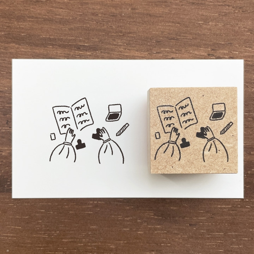 A square Japanese rubber stamp (a-090) featuring the character Marl-chan sitting at a table with stamps and ink pads, illustrating a focused stationery and crafting session. Un tampon en caoutchouc japonais carré (a-090) illustrant le personnage Marl-chan assise à une table avec des tampons et des encreurs, représentant une session de papeterie et d'artisanat concentrée.