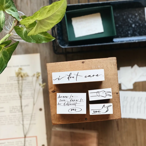 Mr. Eggplants Here Rubber Stamp Set featuring 4 wooden stamps with phrases "i don't care", "here", "yeah", and "brave to love" in a drawer box. Lot de 4 tampons en bois Mr. Eggplants "Here" avec les phrases "i don't care", "here", "yeah", et "brave to love" dans une boîte tiroir.