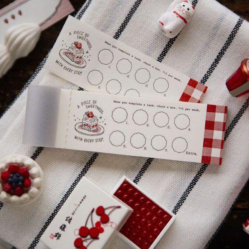 A small, horizontal letterpress notepad with two-color typography and checklist-style designs, featured under a semi-transparent tracing paper cover. Un petit bloc-notes horizontal en typographie (letterpress) avec une mise en page de style "checklist" en deux couleurs, protégé par une couverture en papier calque.