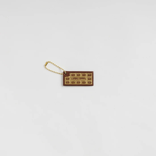 THE SUPERIOR LABOR Leather Tag - Chocolate. A handcrafted leather charm designed to look like a chocolate bar in a silver foil wrapper. Étiquette en cuir THE SUPERIOR LABOR - Chocolat. Un charm artisanal en cuir conçu pour ressembler à une tablette de chocolat dans son emballage argenté.
