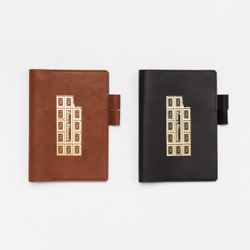 THE SUPERIOR LABOR Note Keeper A5. A handcrafted Tochigi leather notebook cover with industrial-style stamps, shown in Dark Brown and Black. Protège-carnet A5 THE SUPERIOR LABOR. Une couverture de carnet en cuir Tochigi faite à la main avec des tampons industriels, disponible en Marron Foncé et Noir.