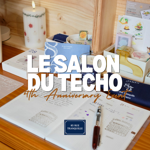 Cozy stationery setup featuring open planners, custom wooden stamps, ink pads, and a TRAVELER'S notebook TOKYO postcard on a wooden workshop table at 85 Rue Tranquille boutique. Espace atelier papeterie chaleureux avec carnets ouverts, tampons personnalisés en bois, encreurs et carte postale TRAVELER'S notebook TOKYO sur une table en bois chez 85 Rue Tranquille.