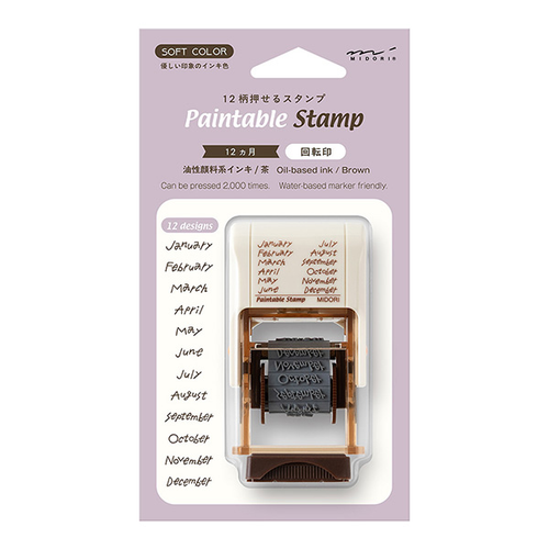 Midori paintable rotating stamp from the Soft Color series in a brown plastic body. Shows the 12 months theme dial making a black ink impression on paper next to water-based markers. / Tampon rotatif à peindre Midori de la série Soft Color avec un corps en plastique marron. Montre le cadran sur le thème des 12 mois imprimant à l'encre noire sur du papier à côté de feutres à base d'eau.