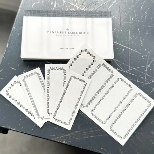OEDA LETTERPRESS 4 Ornament Label Book Frame Black showing a pocket-sized book of perforated paper labels with ornate black printed frames. / Carnet d'étiquettes OEDA LETTERPRESS 4 Ornament Frame Noir présentant un petit carnet d'étiquettes en papier perforé à cadres noirs ornementaux.