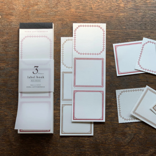 OEDA LETTERPRESS 3 Pattern Label Book Red & Bronze displaying perforated square paper labels with intricate red and metallic bronze printed frames. / Carnet d'étiquettes OEDA LETTERPRESS 3 Pattern Rouge & Bronze présentant des étiquettes carrées en papier perforé imprimées de cadres rouges et bronze.