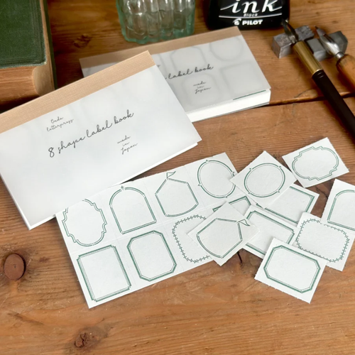 OEDA LETTERPRESS 8 Shape Label Book Limited Edition Frame Green showing a pocket-sized book with tiny perforated green frame labels. / Carnet d'étiquettes OEDA LETTERPRESS 8 Shape Édition Limitée Frame Vert présentant un petit carnet d'étiquettes perforées en papier vert à cadres ornementaux.