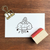 A square Japanese rubber stamp (a-089) featuring the character Marl-chan sitting and using a small rubber stamp on a piece of paper, celebrating the joy of stamping and crafting. Un tampon en caoutchouc japonais carré (a-089) illustrant le personnage Marl-chan assise en train d'utiliser un petit tampon sur une feuille de papier, célébrant la joie de l'estampage et de l'artisanat.