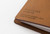TRAVELER'S Notebook Starter Kit Regular Size in Camel. A warm tan leather cover with a blank refill, green spare elastic, and a cotton bag. Kit de démarrage TRAVELER'S Notebook Taille Régulière en Camel. Une couverture en cuir havane avec une recharge vierge et un élastique de rechange vert.