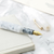 attaché Marbled Fountain Pen in White, featuring a pearlescent white marbled acrylic body and a 24K gold-plated fine nib. Stylo-plume attaché Marbled en blanc, avec un corps en acrylique marbré blanc perlé et une plume fine plaquée or 24 carats.