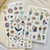 Pion Washi Sticker - Artsy. A sheet of premium die-cut washi stickers featuring delicate illustrations of stylish artsy girls in literary and cozy scenes, on textured washi paper. Autocollant washi Pion - Artsy. Une feuille d'autocollants washi découpés premium avec de délicates illustrations de filles artistiques stylées dans des scènes littéraires et cosy, sur papier washi texturé.