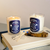 85 Rue Tranquille x ISSMEN Journaling Ritual Set including a canvas tote bag, a scented candle, a blue-tipped matchbox, and an art postcard. / Coffret Rituel de Journaling 85 Rue Tranquille x ISSMEN comprenant un sac cabas en toile, une bougie parfumée, une boîte d'allumettes à bout bleu et une carte postale artistique.