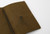 TRAVELER'S Notebook Starter Kit Passport Size in Olive. A forest-green leather cover with a blank refill, blue-gray spare elastic, and a cotton bag. Kit de démarrage TRAVELER'S Notebook Taille Passeport en Olive. Une couverture en cuir vert forêt avec une recharge vierge et un élastique de rechange bleu-gris.