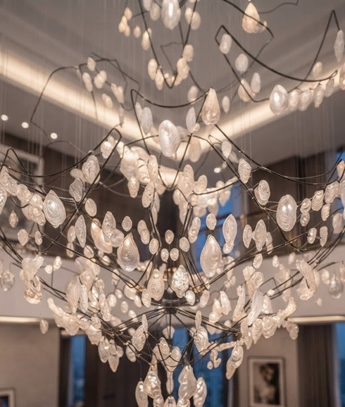 The NORLO chandelier is showcased cleanly against a neutral studio backdrop, highlighting its intricate dark metal frame and suspended, organic glass elements. This product photoshoot view emphasizes the modern craftsmanship of the floating glass pieces and the sophisticated multi-string design.