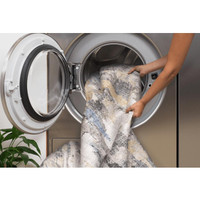 Hands placing an ivory and blue abstract washable rug into a front-loading washing machine.