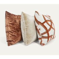 Three luxury throw pillows: a textured copper velvet, a cream velvet with piping, and a white with copper abstract design.