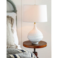 Riviera Table Lamp with a matte white, textured base and a white linen shade on a wooden nightstand.