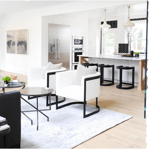 Modern open-concept living room with white armchairs, black counter stools, and light wood floors.