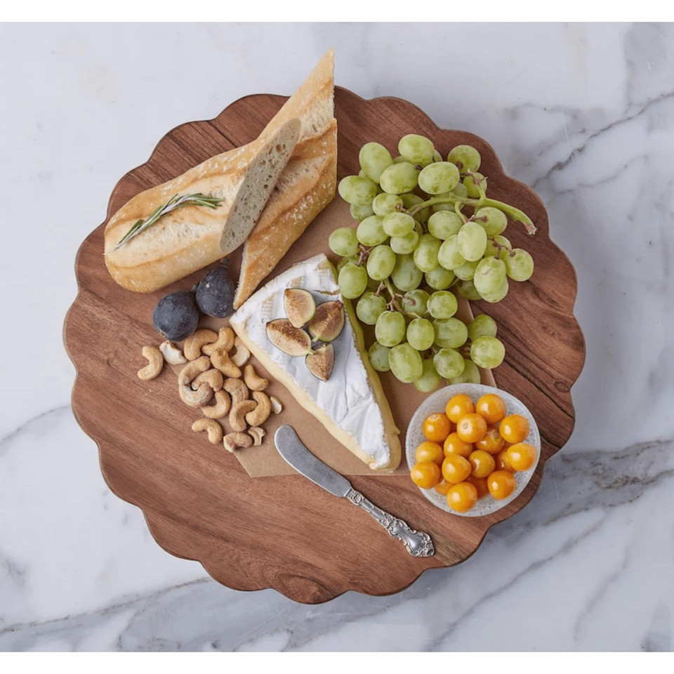 Round scalloped acacia wood charcuterie board with brie, grapes, bread, figs, cashews, and ground cherries.