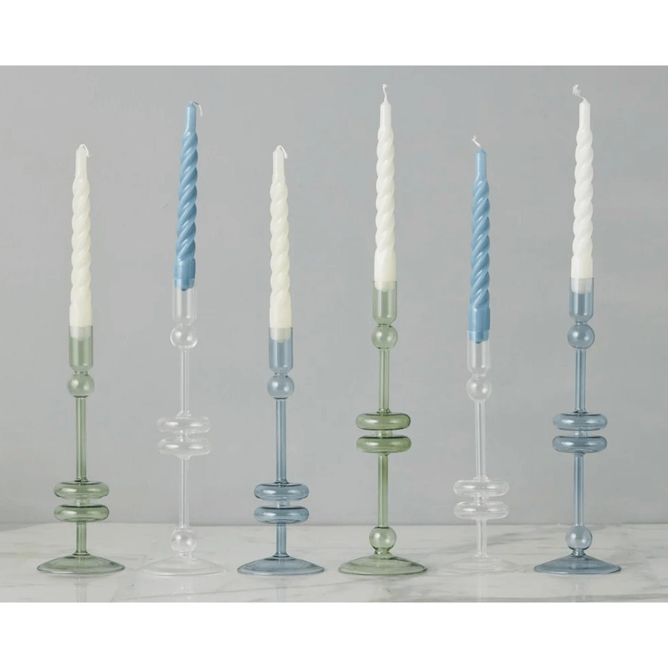 Six elegant glass candlesticks in varying heights and shades of denim blue, clear, and sage green, holding white and blue taper candles.