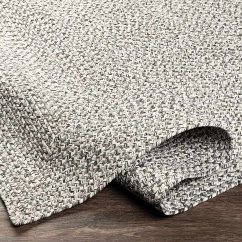 Gray braided polypropylene outdoor rug with a no-pile texture, partially rolled on a wood floor.