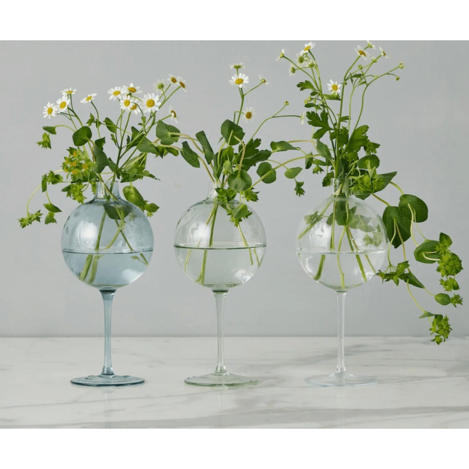 Three clear glass wine glass vases, one blue, one light green, and one clear, each holding small white flowers.