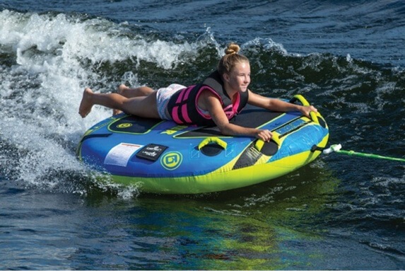 Girl riding blue-green towable boat tube across wake in pink life vest