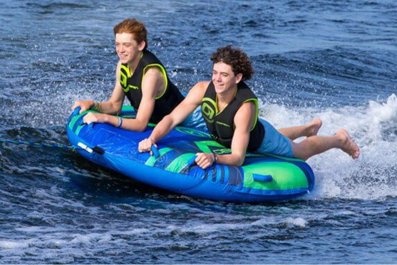 Two teens enjoying blue inflatable water tube skimming across lake