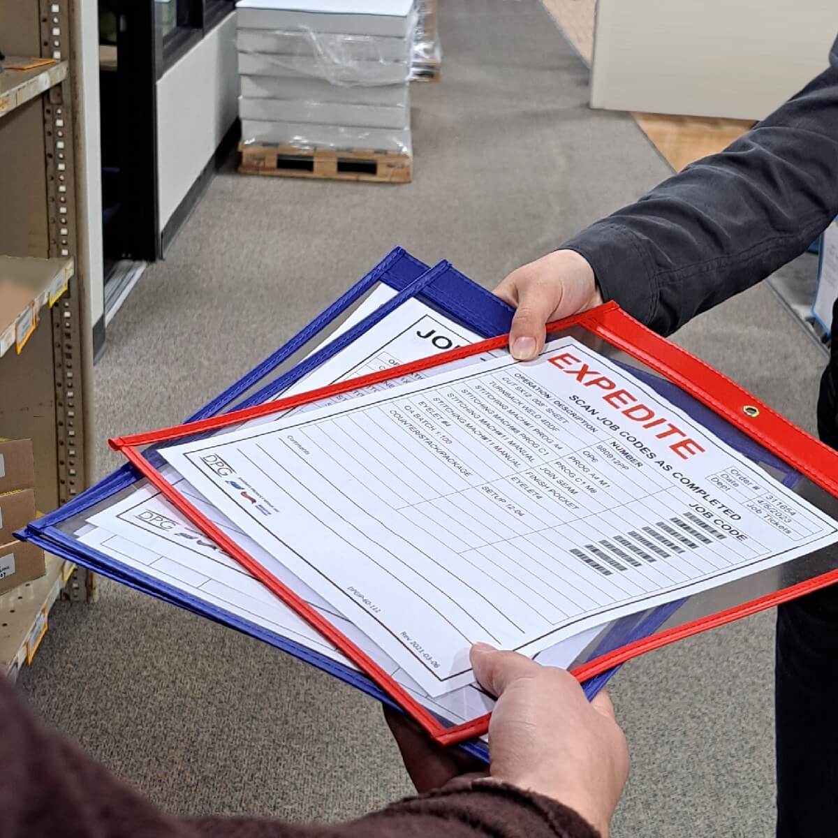 Worker handing off production paperwork in a warehouse aisle using heavy-duty job ticket holders for shop floor document control