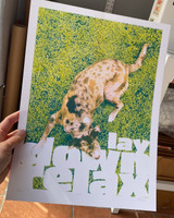 product image of a human hand holding the art print in their left hand. Art print features a dog on a green grass background with white text saying "lay down relax" along the bottom 1/3rd.