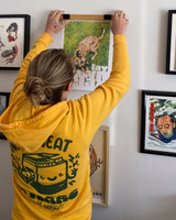 Image of a person from behind hanging a frame on the wall wearing the product hoodie [zip option] hoodie is golden yellow with a hood and dark green ink printed on back. image printed on back is a printer with the words "ink, sweat, and tears": & "split arrow" underneath
