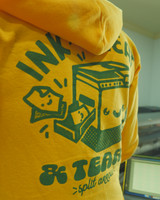 Zoomed in image of Image of a person from behind at a risograph printer  wearing the product hoodie [zip option] hoodie is golden yellow with a hood and dark green ink printed on back. image printed on back is cropped in photo with a portion of text being legible.