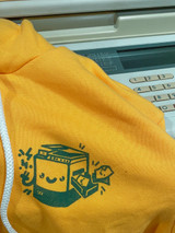 Image of the product hoodie unworn. Hoodie is gold yellow with a green ink image of a printer with a face. Hoodie is laying on a 90's risograph