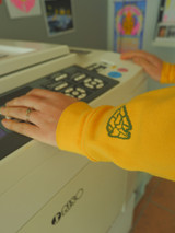 image of an arm controlling a risograph printer with art prints hanging on walls in background. Person is wearing the product hoodie [zip option] image shows the Split Arrow Print House logo in dark green printed on the bottom of the sleeve