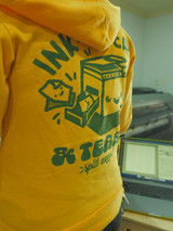 Zoomed in image of Image of a person from behind at a risograph printer  wearing the product hoodie [zip option] hoodie is golden yellow with a hood and dark green ink printed on back. image printed on back is cropped in photo with a portion of text being legible.