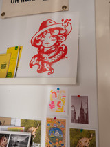 Product art print featuring a lady head with a hat and flag scarf printed in red ink on white paper on a shelf on a wall surrounded by other art and books