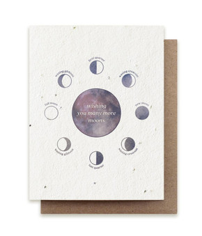 A plantable birthday card embedded with herb seeds and a purple watercolor design of moon phases saying "wishing you many more moons" with a kraft envelope on a white background